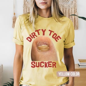 Pu&ograve; includere: T-shirt gialla con la scritta "DIRTY TOE SUCKER" in lettere rosse. La maglietta presenta un'immagine ravvicinata di un dito del piede con una condizione dell'unghia. Le parole "YELLOW COLOR" sono in basso sulla maglietta.