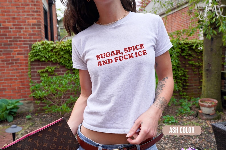 May include: A light grey t-shirt with the red text "SUGAR, SPICE AND FUCK ICE". The shirt is being worn. The background features a brick wall and some green foliage.