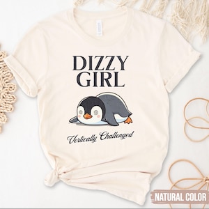 Puede incluir: Una camiseta beige con un gráfico negro de un pingüino mareado y el texto "Dizzy Girl Vertically Challenged".
