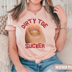 Pu&ograve; includere: T-shirt color pesca con la scritta "DIRTY TOE SUCKER" in rosso. La grafica presenta un primo piano di un dito del piede con un'infezione fungina. La maglietta &egrave; realizzata in materiale morbido ed &egrave; adatta per un abbigliamento casual.