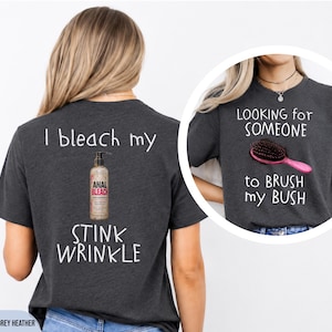 May include: A dark grey heather t-shirt featuring white text on the back that reads I bleach my stink wrinkle with a bottle graphic, and front text that reads Looking for someone to brush my bush with a pink hairbrush graphic.