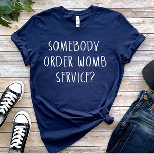 Op de afbeelding: Een marineblauw T-shirt met witte tekst die "Heeft iemand een baarmoederdienst besteld?" luidt. Het shirt ligt plat op een houten oppervlak met een paar zwarte sneakers, een blauwe jeans en een zwarte baseballpet.