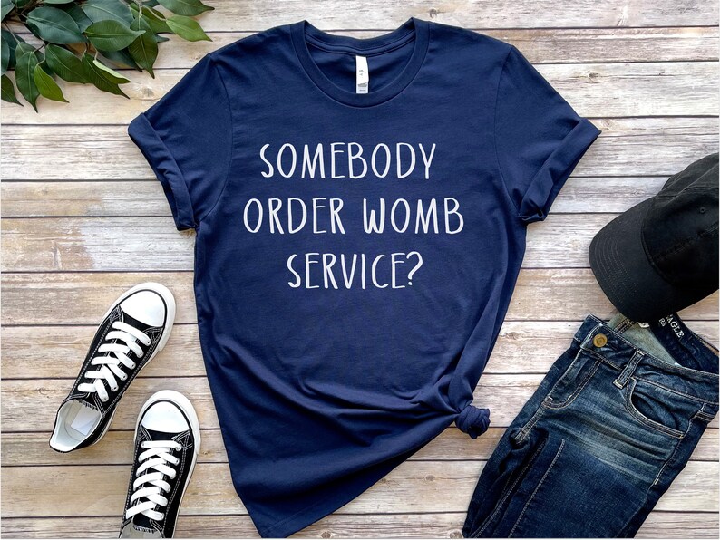Op de afbeelding: Een marineblauw T-shirt met witte tekst die "Heeft iemand een baarmoederdienst besteld?" luidt. Het shirt ligt plat op een houten oppervlak met een paar zwarte sneakers, een blauwe jeans en een zwarte baseballpet.
