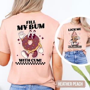 May include: Heather peach-colored t-shirt with a cartoon donut graphic on the back, featuring the text "FILL MY BUM WITH CUM!". The front of the shirt has a cartoon ice cream cone with the text "LICK ME" and "'TIL ICE CREAM".