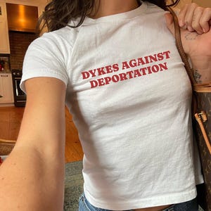 May include: White t-shirt with the text "DYKES AGAINST DEPORTATION" in red block letters. The shirt is a crew neck style. The person is also wearing blue jeans.