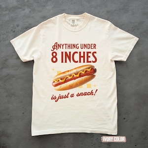 May include: Ivory-colored t-shirt with the text "ANYTHING UNDER 8 INCHES is just a snack!" and a graphic of a hot dog with mustard. The shirt is a casual wear item.