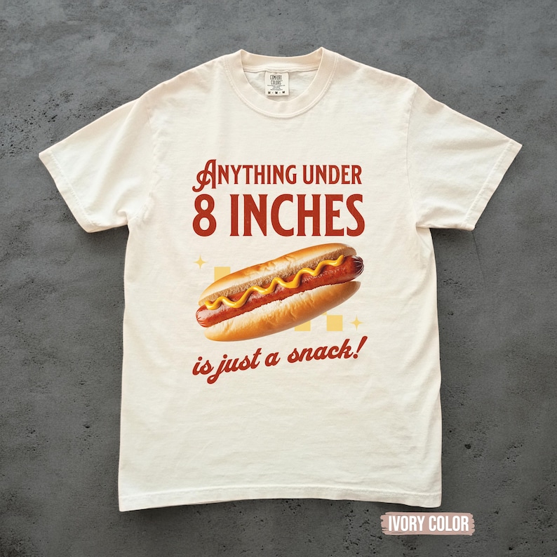 May include: Ivory-colored t-shirt with the text "ANYTHING UNDER 8 INCHES is just a snack!" and a graphic of a hot dog with mustard. The shirt is a casual wear item.