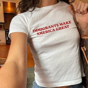 Puede incluir: Camiseta blanca con el texto rojo "IMMIGRANTS MAKE AMERICA GREAT". La camiseta es de estilo ajustado y de manga corta. La persona también lleva vaqueros azules y sostiene un bolso marrón.