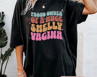 May include: Black heather oversized t-shirt with the text "PROUD OWNER OF A HUGE SMELLY VAGINA" in colorful retro font. The shirt has short sleeves and a crew neck. The text is in shades of blue, orange, yellow, and pink.