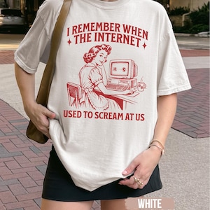 May include: An off-white short-sleeve t-shirt featuring a red graphic of a person sitting at a vintage computer. The text reads, I REMEMBER WHEN THE INTERNET USED TO SCREAM AT US. The design includes small star accents and a coffee mug on the desk.
