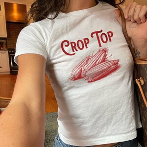 Op de afbeelding: Witte crop top met de rode tekst "Crop Top" en een rode illustratie van drie maïskolven. De top met korte mouwen is gemaakt van zachte stof.