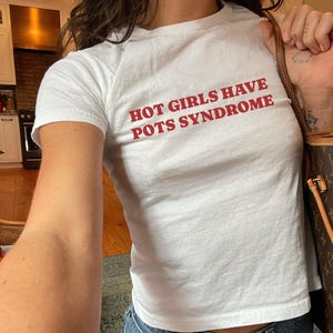 Puede incluir: Camiseta blanca con el texto rojo "HOT GIRLS HAVE POTS SYNDROME". La camiseta es de cuello redondo y parece estar hecha de un material suave. La camiseta se combina con unos vaqueros azules.