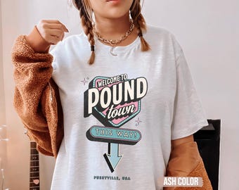 Puede incluir: Camiseta color ceniza con un gráfico de letrero retro que dice "WELCOME TO POUND TOWN THIS WAY!" en negro, rosa y turquesa. El texto "PUSSYVILLE, USA" está impreso debajo. La camiseta tiene mangas cortas.