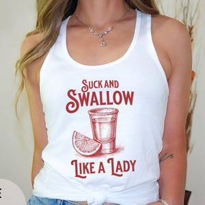May include: White vest top with a red graphic that reads "Suck and Swallow Like a Lady." The design features a shot glass and a lemon slice. The top is a casual, sleeveless style.