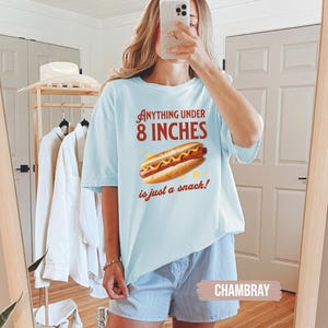 May include: Light blue t-shirt with the text "Anything Under 8 Inches is just a snack!" and a graphic of a hot dog. The shirt is paired with blue and white striped shorts.