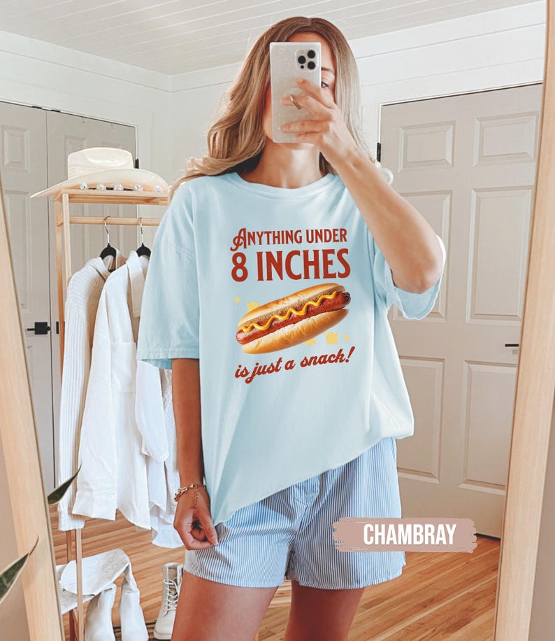 May include: Light blue t-shirt with the text "Anything Under 8 Inches is just a snack!" and a graphic of a hot dog. The shirt is paired with blue and white striped shorts.