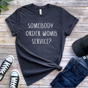Op de afbeelding: Een donkergrijs T-shirt met de tekst "SOMEBODY ORDER WOMB SERVICE?" in wit gedrukt. Het shirt ligt plat op een houten oppervlak met een paar zwarte sneakers, een zwarte baseballpet en een blauwe jeans.
