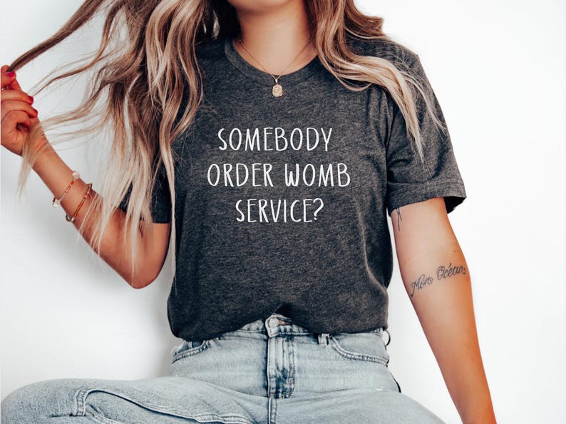 May include: A dark grey t-shirt with the text "Somebody order womb service?" printed in white.