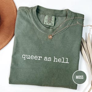 Pode incluir: Uma camiseta verde musgo com a frase "queer as hell" impressa em branco. A camiseta tem gola redonda e mangas curtas. Um colar de ouro com um pingente está drapeado sobre a camiseta. Um chapéu marrom é visível no canto superior esquerdo.
