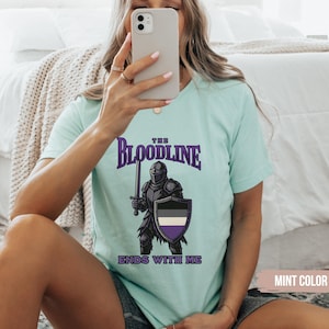 Puede incluir: Una camiseta azul claro con un gr&aacute;fico de un caballero sosteniendo una espada y un escudo. El escudo tiene un dise&ntilde;o negro, blanco y morado. El texto en la camiseta dice "The Bloodline Ends With Me".