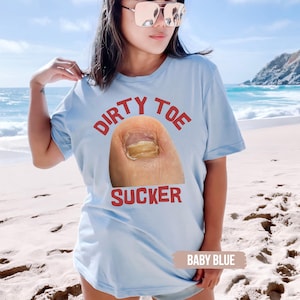 Pu&ograve; includere: T-shirt azzurro con la scritta rossa "DIRTY TOE SUCKER" e una grafica di un dito del piede con un'unghia gialla. La maglietta &egrave; di colore azzurro.