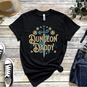 May include: Black t-shirt with a graphic of a sword and dice with the text "Dungeon Daddy".