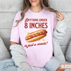 May include: Light pink t-shirt with the text "Anything under 8 INCHES is just a snack!" and a graphic of a hot dog. The hot dog is on a bun with mustard. The shirt is a blossom color.