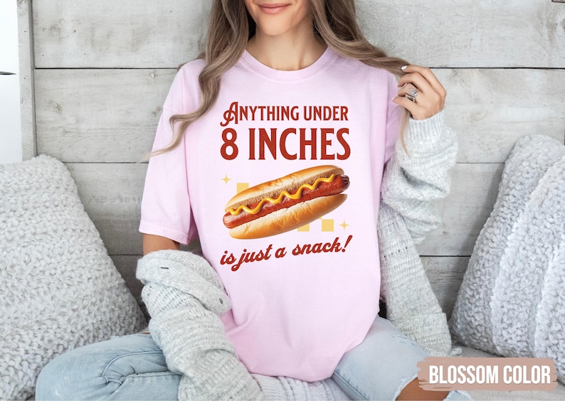 May include: Light pink t-shirt with the text "Anything under 8 INCHES is just a snack!" and a graphic of a hot dog. The hot dog is on a bun with mustard. The shirt is a blossom color.
