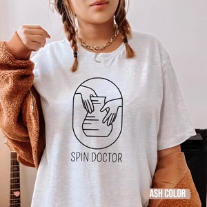 Pode incluir: Camiseta cinza claro com um desenho de linha preta de mãos moldando um vaso. O texto "SPIN DOCTOR" está abaixo do desenho. Parte de uma coleção de roupas.