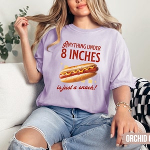 May include: Orchid-colored t-shirt with the text "Anything under 8 inches is just a snack!" and a graphic of a hot dog. The shirt is a comfortable, casual style.