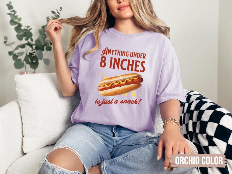 May include: Orchid-colored t-shirt with the text "Anything under 8 inches is just a snack!" and a graphic of a hot dog. The shirt is a comfortable, casual style.
