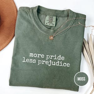 May include: A moss green t-shirt with the text "more pride less prejudice" in white. A gold necklace with a pendant rests on the shirt. The shirt is folded and has a tag that says "Comfort Colors".
