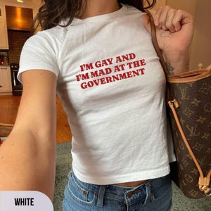 May include: White crew neck t-shirt with the red text "I'M GAY AND I'M MAD AT THE GOVERNMENT". The t-shirt is worn with blue jeans. A brown handbag with a pattern is also visible in the image.