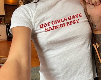May include: White t-shirt with the red text "HOT GIRLS HAVE NARCOLEPSY". The shirt is short-sleeved and appears to be made of a soft, lightweight material. The text is centered on the front of the shirt.