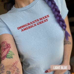 May include: Light blue t-shirt with the text "IMMIGRANTS MAKE AMERICA GREAT" in red. The shirt is a crew neck style and appears to be made of a soft material. The image also shows a purple braid and a rose tattoo.