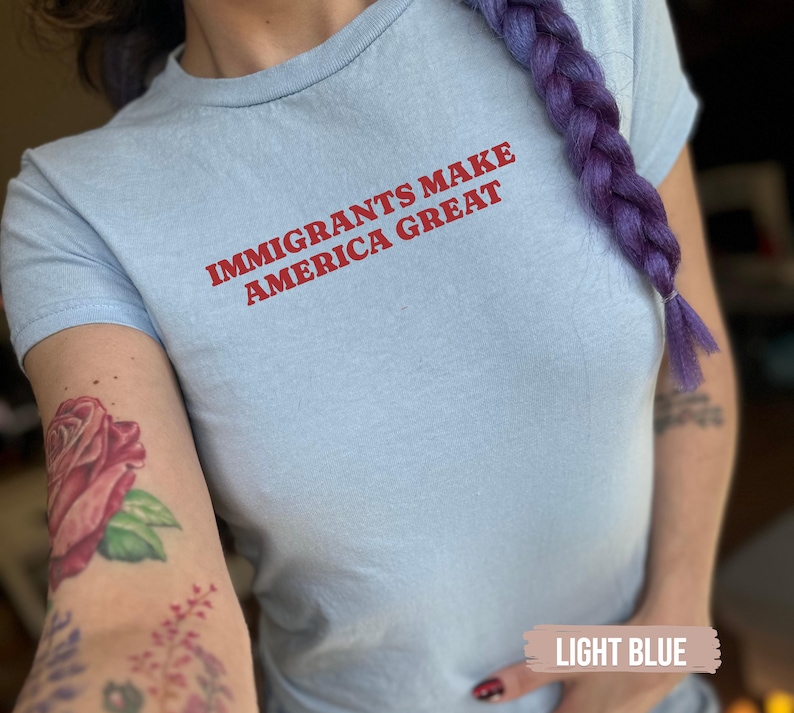 May include: Light blue t-shirt with the text "IMMIGRANTS MAKE AMERICA GREAT" in red. The shirt is a crew neck style and appears to be made of a soft material. The image also shows a purple braid and a rose tattoo.