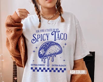 May include: Ash-colored t-shirt with a blue graphic of a taco and the text "ASK FOR A TASTE OF MY SPICY TACO." The design includes the phrases "EXTRA CHEESY" and "EXTRA SLEAZY" below the taco illustration.