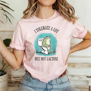 May include: A pink t-shirt with a graphic of a frog sitting on a toilet. The text on the shirt reads "I tolerate a lot but not lactose".