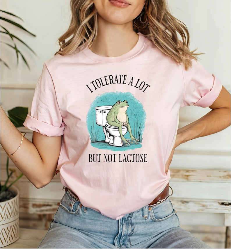 May include: A pink t-shirt with a graphic of a frog sitting on a toilet. The text on the shirt reads "I tolerate a lot but not lactose".