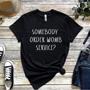 Op de afbeelding: Zwart T-shirt met witte tekst die "Heeft iemand een baarmoederdienst besteld?" zegt.