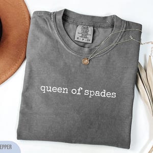 Może przedstawiać: Koszulka z krótkim rękawem w kolorze pieprzowym z okrągłym dekoltem. Na piersi znajduje się biały napis "queen of spades". Wewnątrz przy kołnierzu widoczna jest metka marki Comfort Colors wskazująca na rozmiar S.