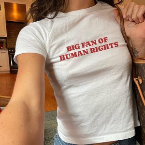 Op de afbeelding: Wit T-shirt met de rode tekst "BIG FAN OF HUMAN RIGHTS". Het shirt heeft een ronde hals en lijkt gemaakt van een zachte, lichte stof. De persoon draagt een blauwe spijkerbroek.