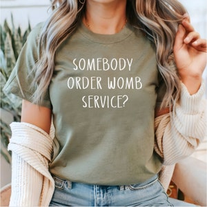 Op de afbeelding: Een groen T-shirt met de tekst "Somebody order womb service?" in wit.