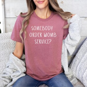 Op de afbeelding: Een vrouw draagt een mauve t-shirt met de tekst "Somebody order womb service?" in wit.
