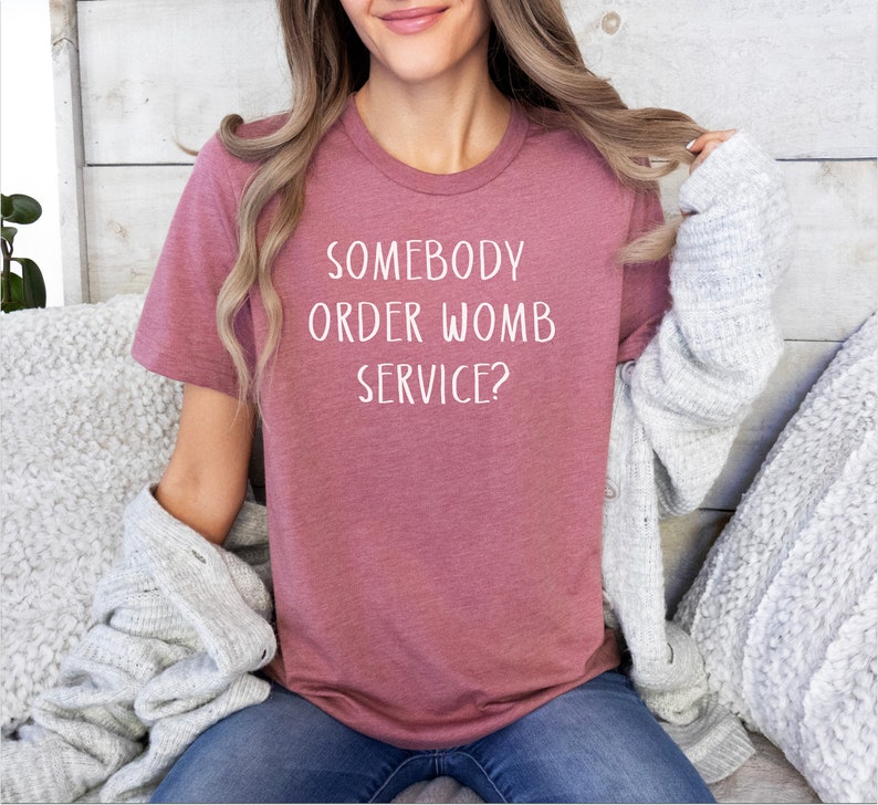Op de afbeelding: Een vrouw draagt een mauve t-shirt met de tekst "Somebody order womb service?" in wit.