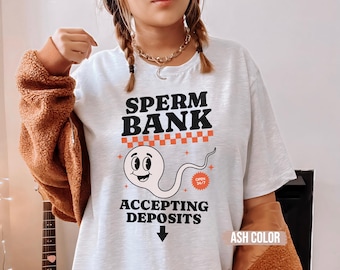 Puede incluir: Camiseta gris ceniza con el texto "SPERM BANK" en negro y naranja, con un espermatozoide de dibujos animados y las palabras "ACCEPTING DEPOSITS". La camiseta también tiene un gráfico "OPEN 24/7".