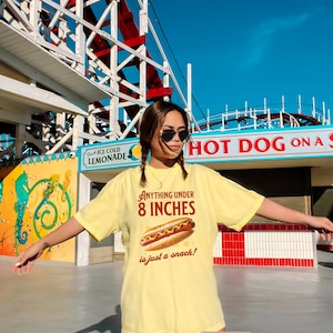 May include: A light yellow t-shirt with the text "Anything under 8 inches is just a snack!" and a graphic of a hot dog. The shirt is labeled "Banana Color". The background includes a roller coaster and a hot dog stand.