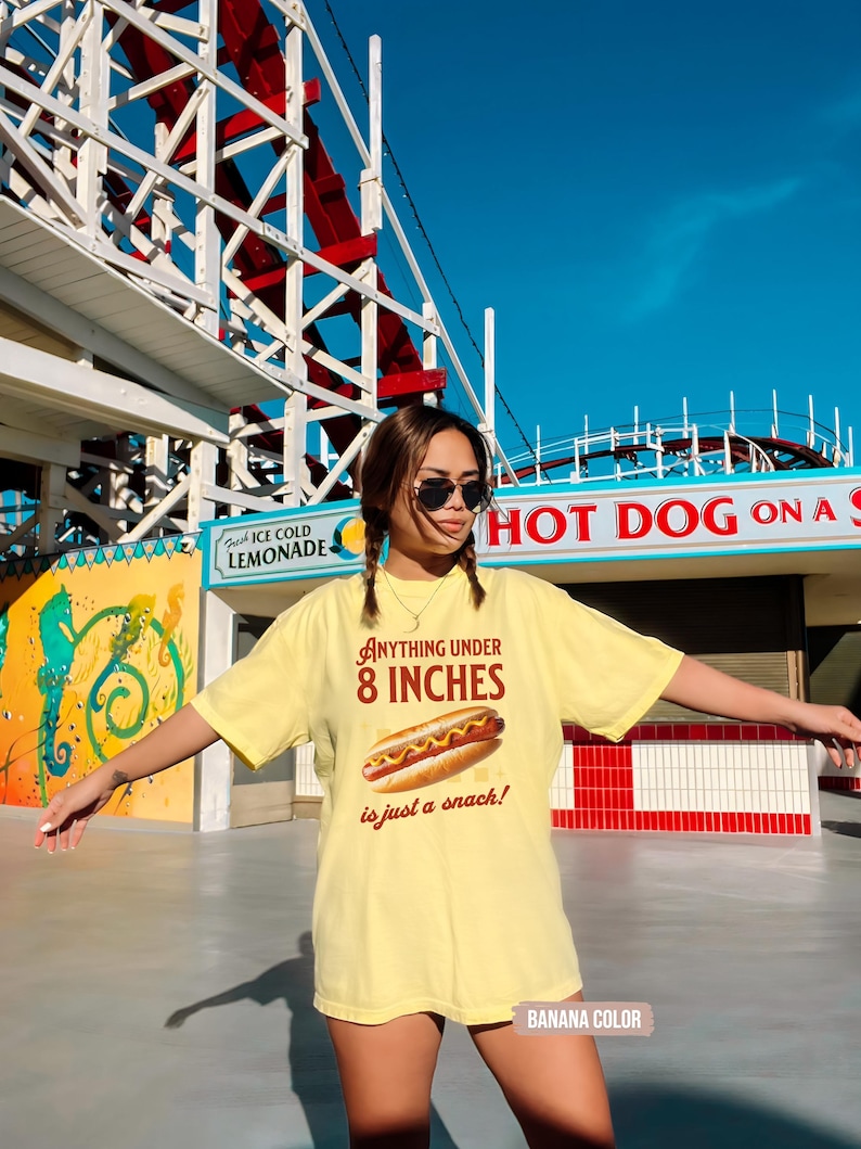 May include: A light yellow t-shirt with the text "Anything under 8 inches is just a snack!" and a graphic of a hot dog. The shirt is labeled "Banana Color". The background includes a roller coaster and a hot dog stand.