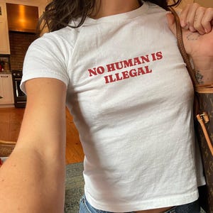 May include: White t-shirt with the red text "NO HUMAN IS ILLEGAL". The short-sleeved shirt is made of a soft material and is paired with blue jeans. A brown handbag is visible in the background.