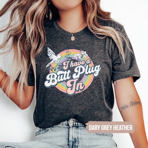 May include: Dark grey heather t-shirt with a colorful graphic. The graphic features a rainbow, flowers, a bird, and the text "I have a Butt Plug In". The shirt is a casual style.